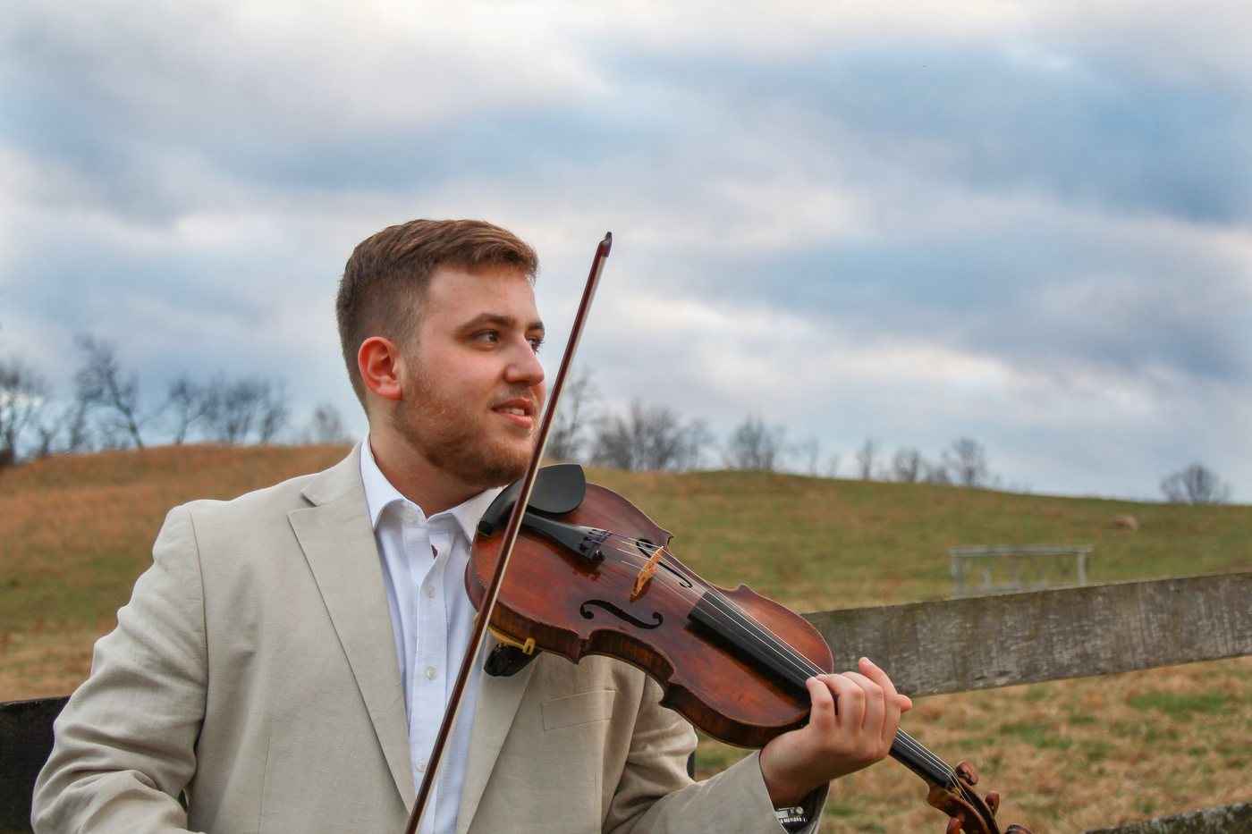 George Pekarsky with his violin outdoors, against an open field and clouded sky