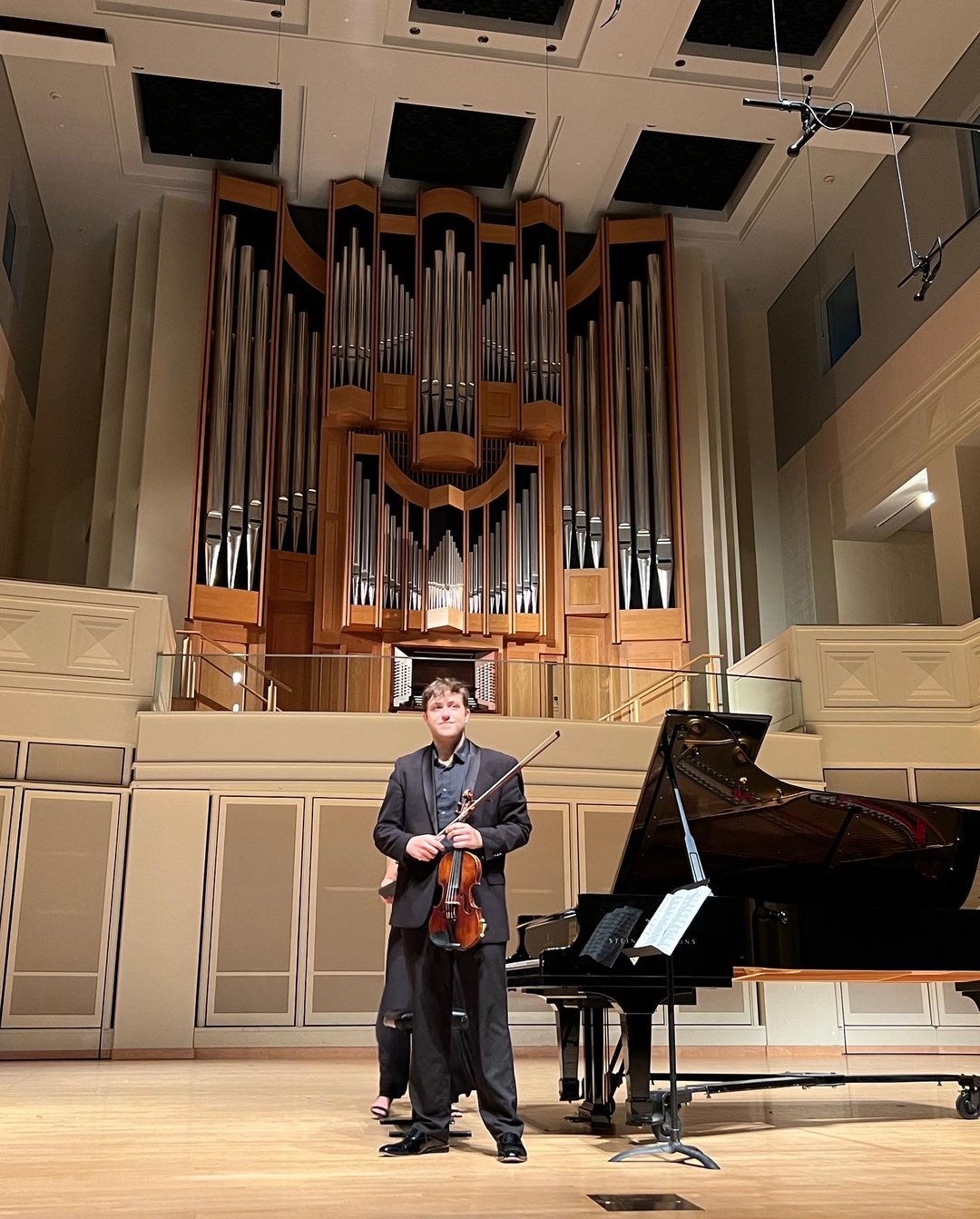 George Pekarsky onstage with his violin beside a grand piano, beneath a large pipe organ